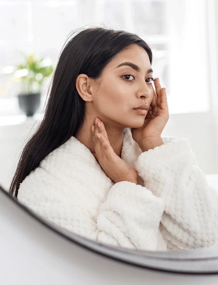 woman looking into the mirror looking young healthy skin aquagold | Geria Dermatology New Jersey