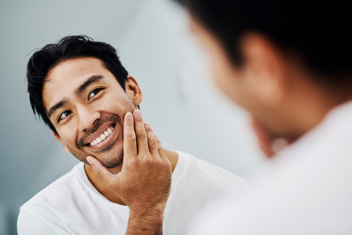Handsome male touching his healthy face in the mirror after a skincare treatment. Confident man feeling fresh and attractive from a beauty routine. Portrait of a guy looking at his skin.